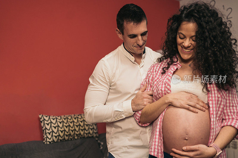 Expectant Heterosexual Couple at Home, Woman Holding Belly, Man Observing from Behind. Pregnant woman holding belly with both hands while her man is behind her observing.